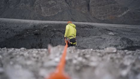 a dynamic shot features a person in bright yellow outdoor gear, seemingly on a rope, positioned against a dark, rugged, and possibly icy or rocky glacial landscape
