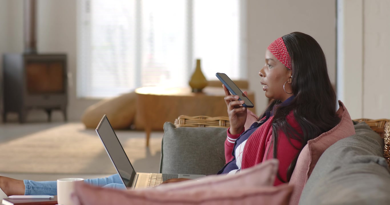 African American woman raising smartphone, speaking on work call while glancing at laptop on sofa