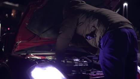 man looking at engine of broken down car with flashlight at night. headlights turned on