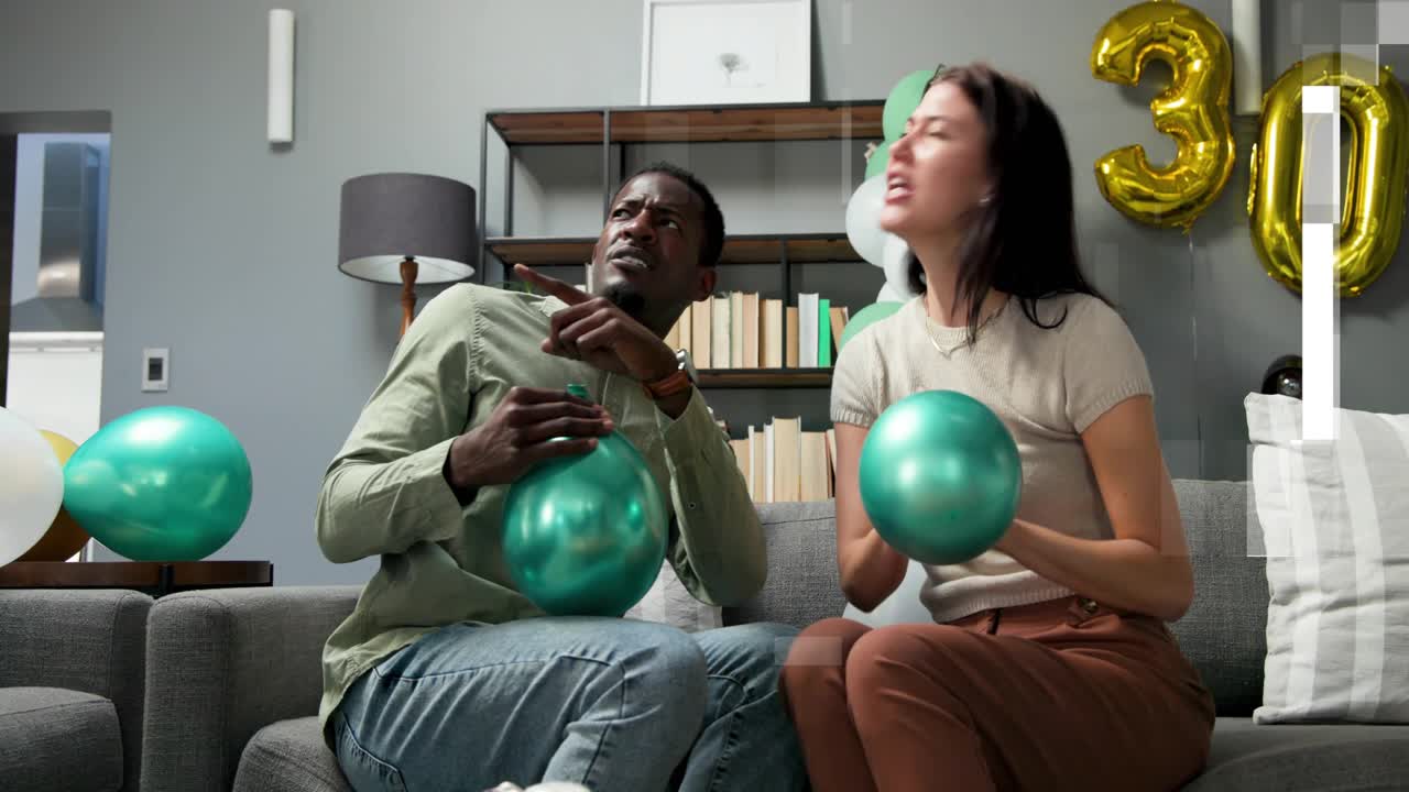 Man pointing upward and woman inflating green foil balloons on sofa, prepping 30th party decor