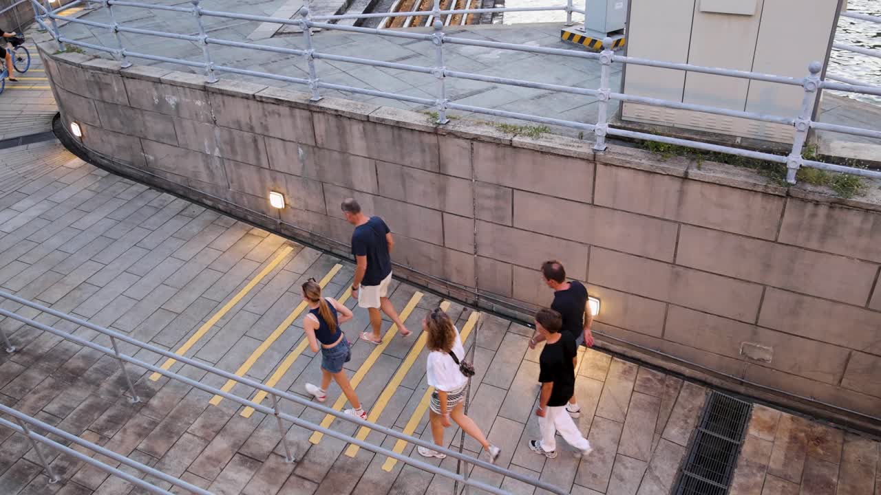 Pedestrians and cyclist move along riverside steps at dusk, captured from elevated, wide-angle perspective