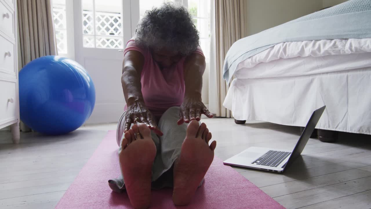 Senior african american woman performing stretching exercise while looking at laptop at home
