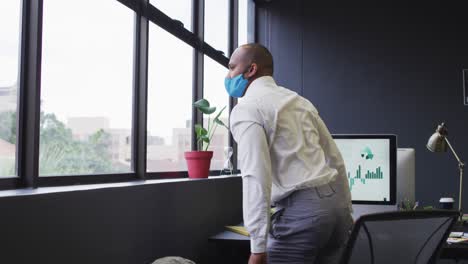 Mixed-race-businessman-wearing-face-mask-standing-looking-out-of-window-in-modern-office