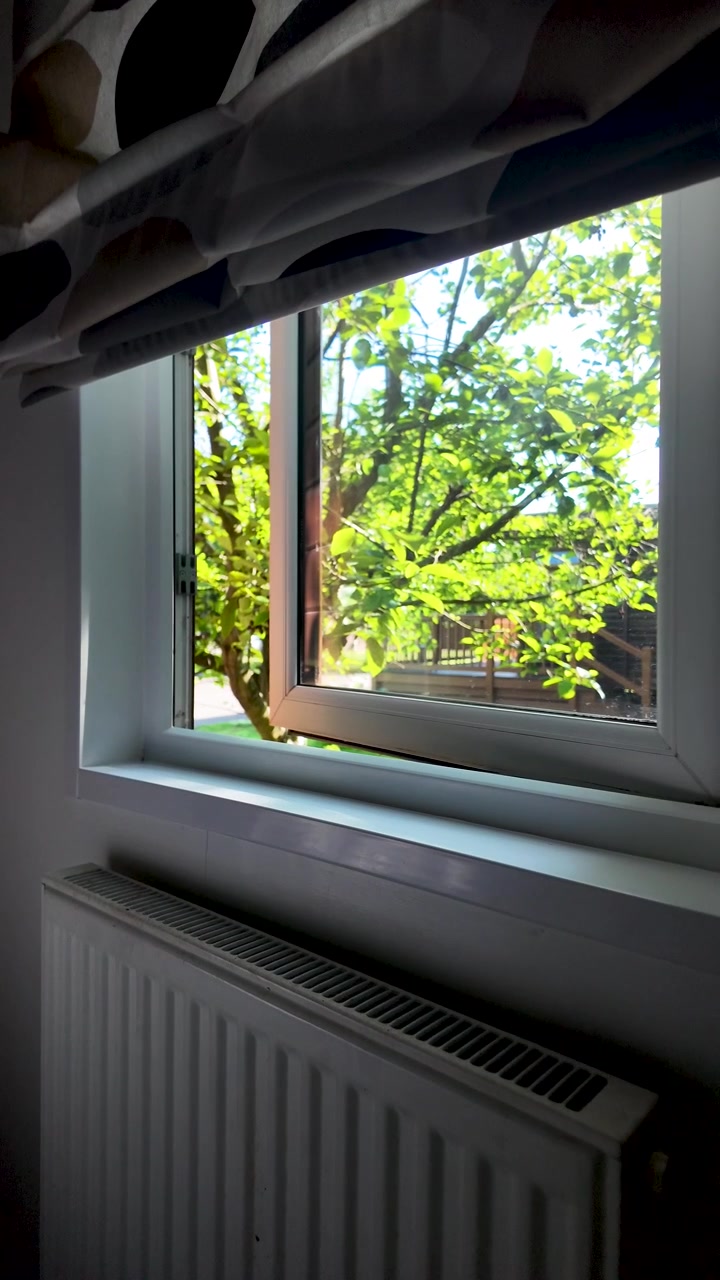 Interior radiator beneath an open window with bright green garden tree outside on a sunny day. vertical video