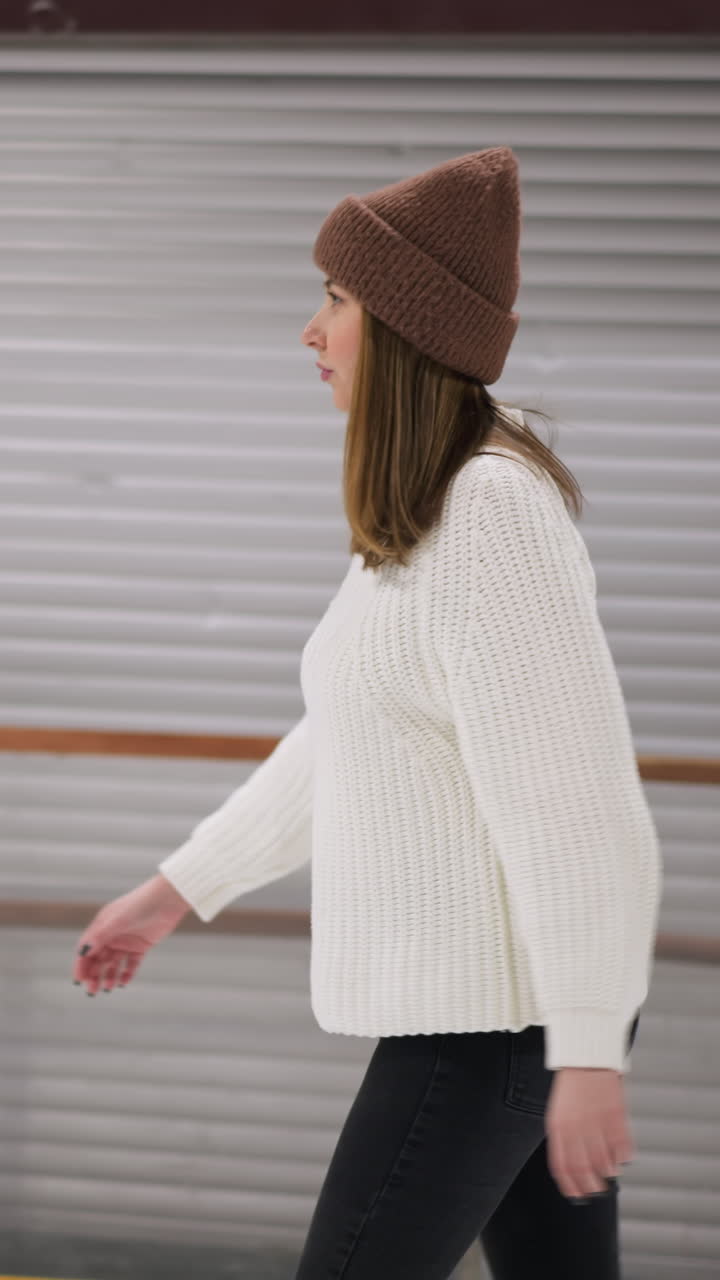 Eine Nahaufnahme einer Frau, die gemütlich auf einer Eisbahn Schlittschuh läuft. Sie trägt einen weißen Pullover und eine braune Mütze, mit einer Ziegelwand und einigen bunten Ballons im Hintergrund.