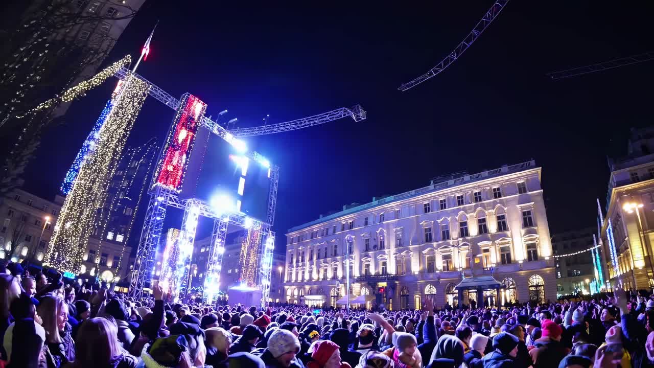 Wide-angle night shot of a vibrant city square, filled with a festive crowd and illuminated screens
