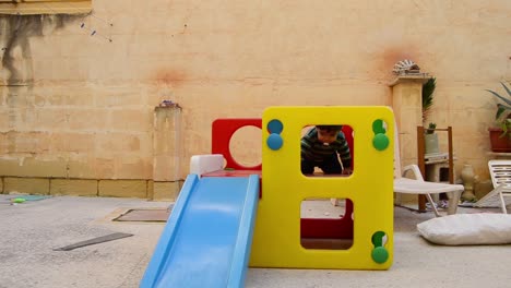 cute two years old boy dirty from the cookies around the mouth sliding down the plastic slide play house in backyard