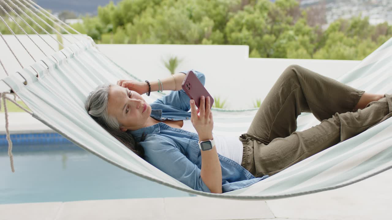 Ældre kvinde ligger i en hvid hængekøje ved siden af poolen, rækker ud efter sin smartphone og scroller.
