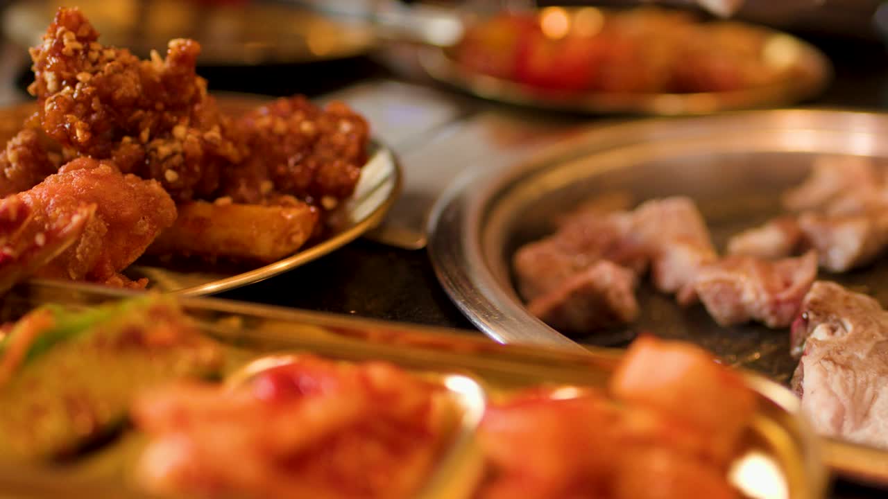 Various meats cook on a tabletop grill, surrounded by side dishes, in warm ambient lighting
