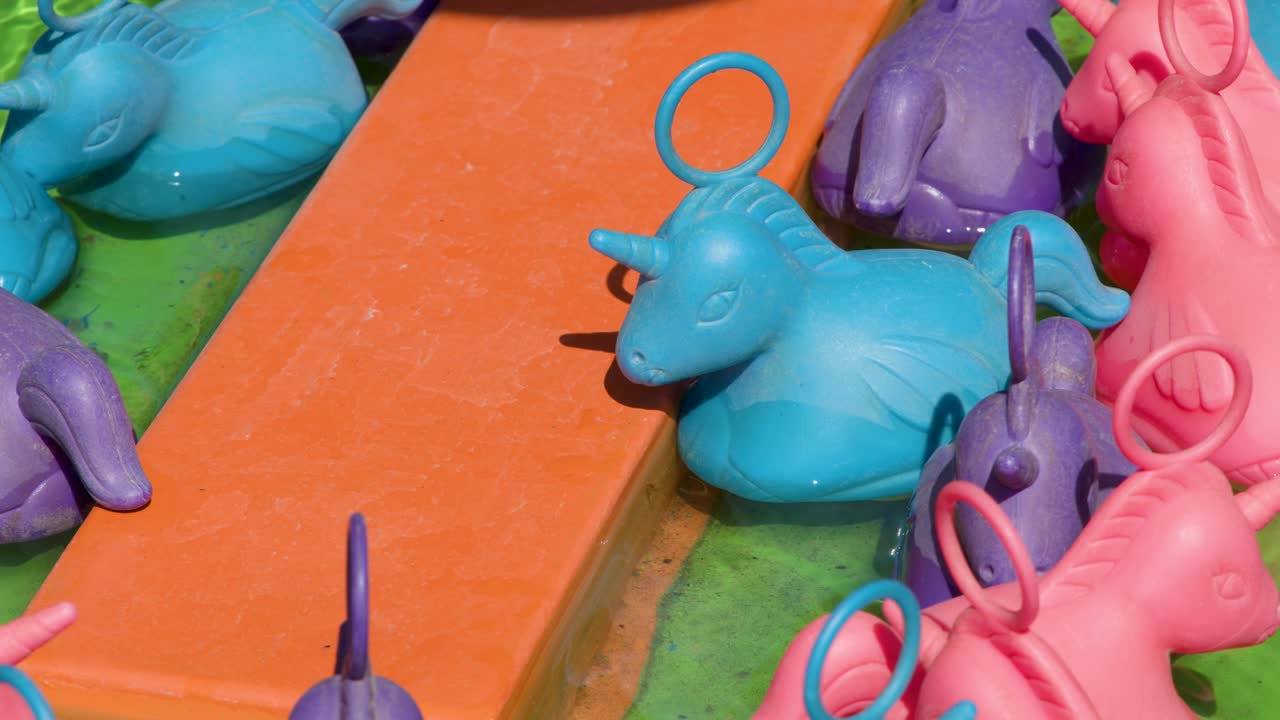 Plastic unicorn toys float in water at a vibrant carnival game, bright daylight, close-up view