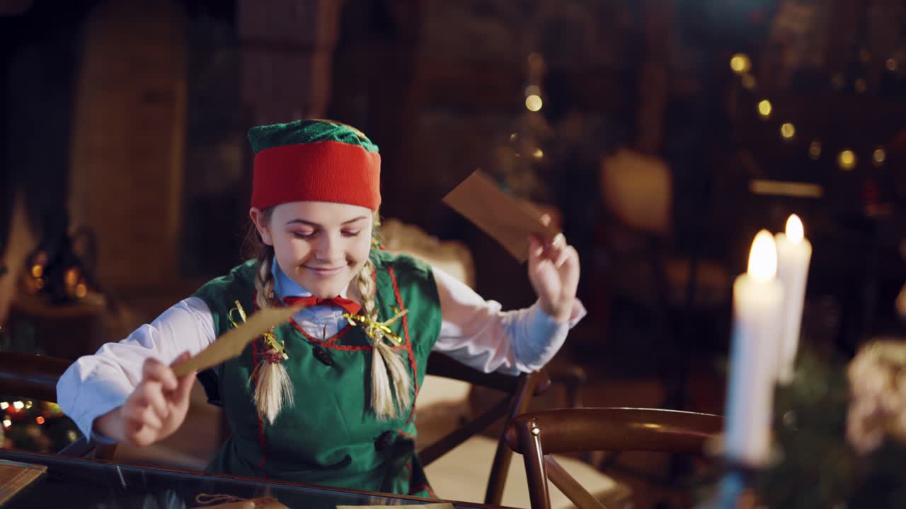 A happy elf is sitting at the table and dancing with two envelopes in her hands on the background of a fireplace and a Christmas tree. Blurred Background