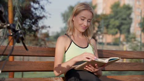 Mujer-Atleta-Feliz-Disfrutando-Leyendo-Un-Libro-Sentado-En-Un-Banco-Al-Aire-Libre