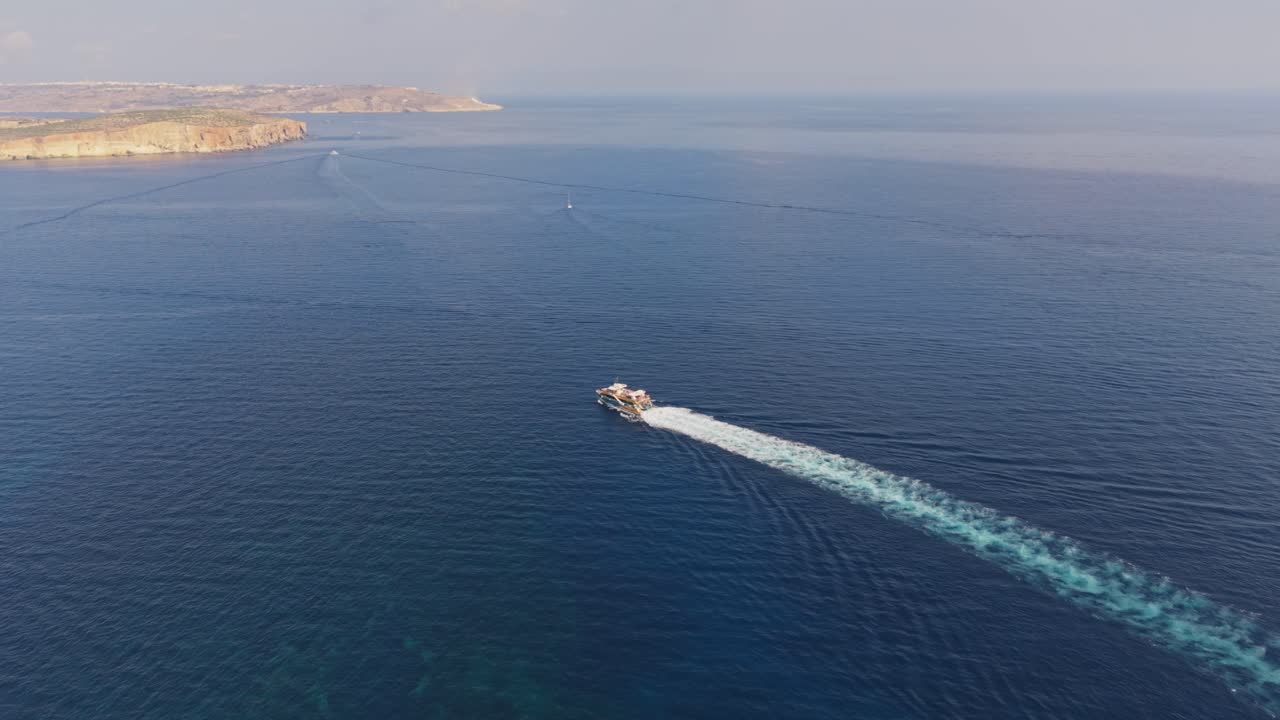 A motorboat cruises across the calm blue sea, leaving a long white trail behind. The distant coastline and cliffs create a serene horizon under a hazy sky