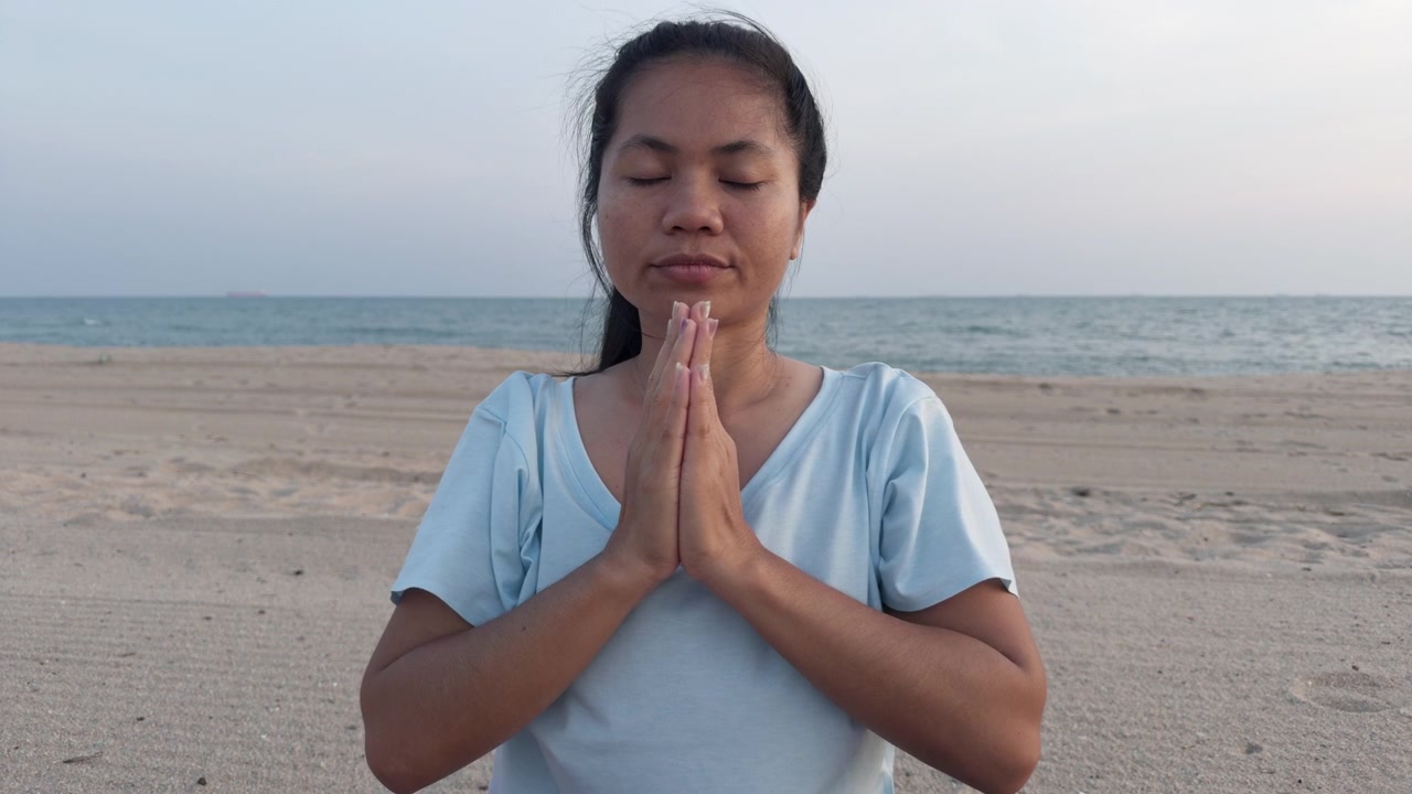 Detailní záběr asijské ženy praktikující jóga meditaci na písečné pláži. Klidné dechové cvičení s modlitebním gestem anjali mudra a zavřenýma očima během poklidného rána u oceánu