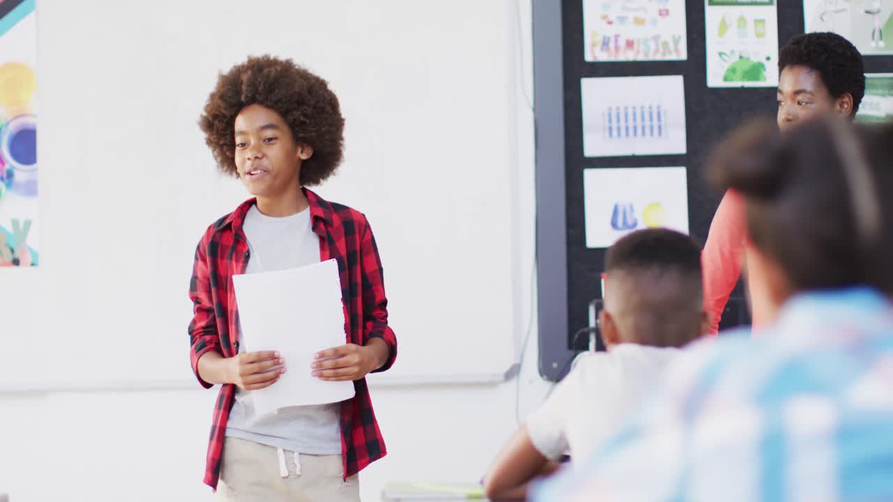 Diverse female teacher and happy schoolchildren at desks reciting in school classroom