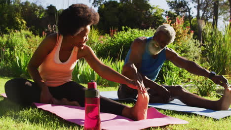 una pareja de ancianos afroamericanos haciendo ejercicio al aire libre sentados estirándose en un jardín soleado