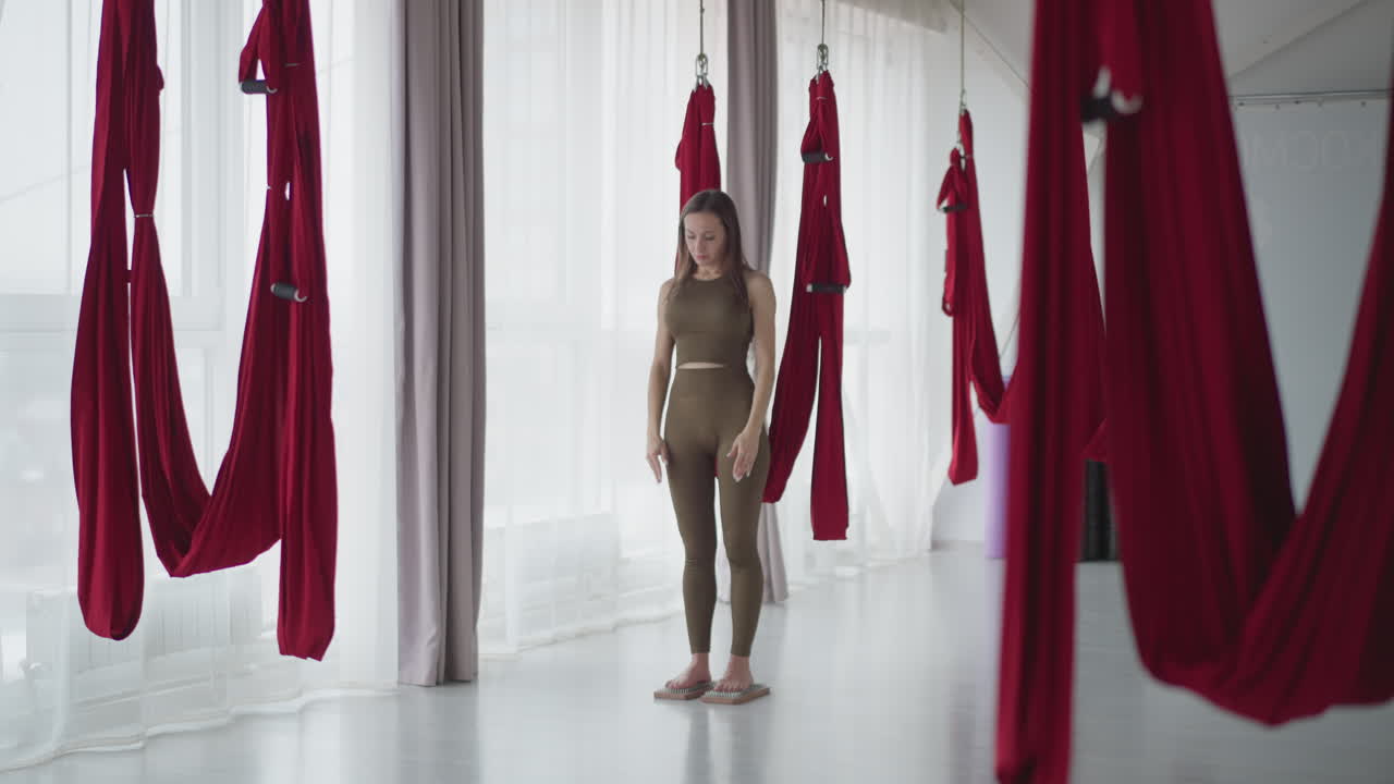 Female athlete practicing quietness barefoot on nail boards in spa like studio surrounded by red hammocks meditating balancing mind body sensory focus under serene ambient peaceful concentration