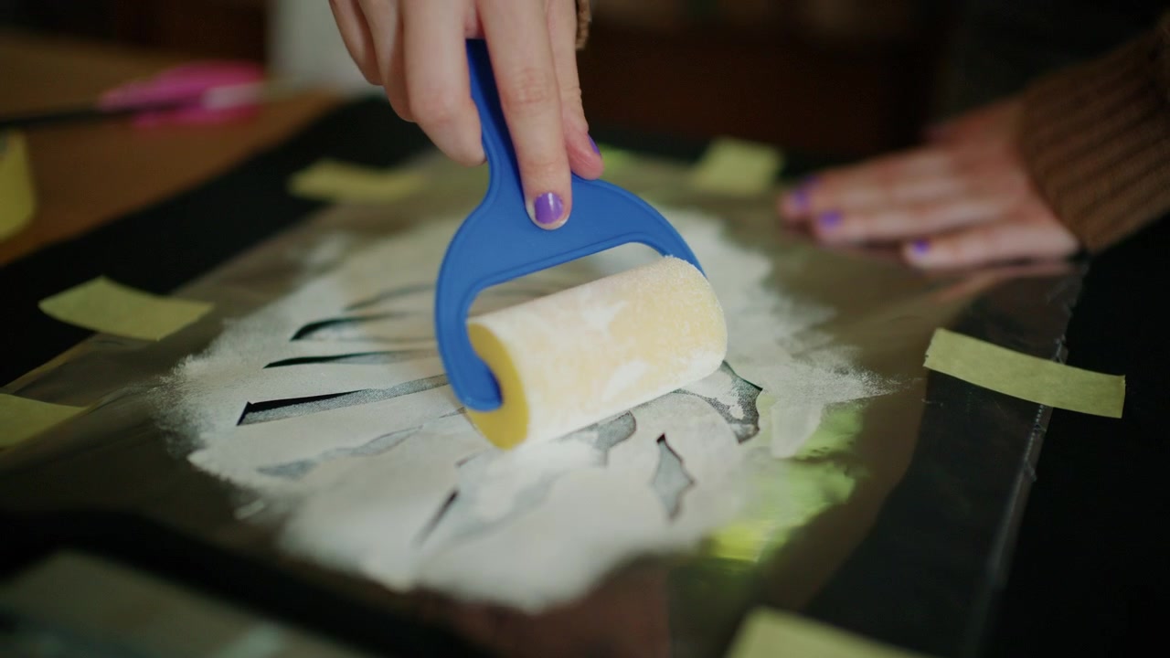 Mani che utilizzano un rullo artigianale per stendere uniformemente materiale adesivo su un disegno a stencil.
