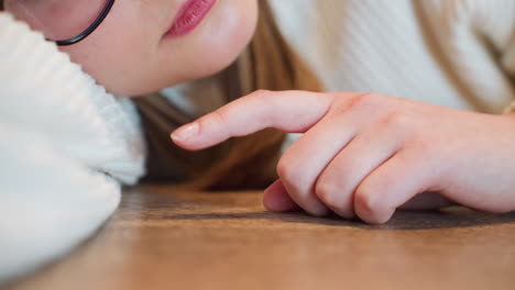 close up of woman lying on wooden table tapping tabletop with finger wearing glasses and sweater showing boredom fatigue and relaxed mood in cozy indoor setting with greenery in background