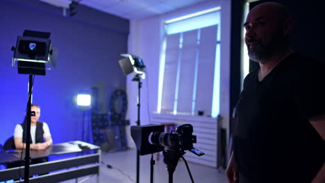 bearded cameraman prepares camera for footage in studio. caucasian female blogger sits at desk at blurred backdrop.