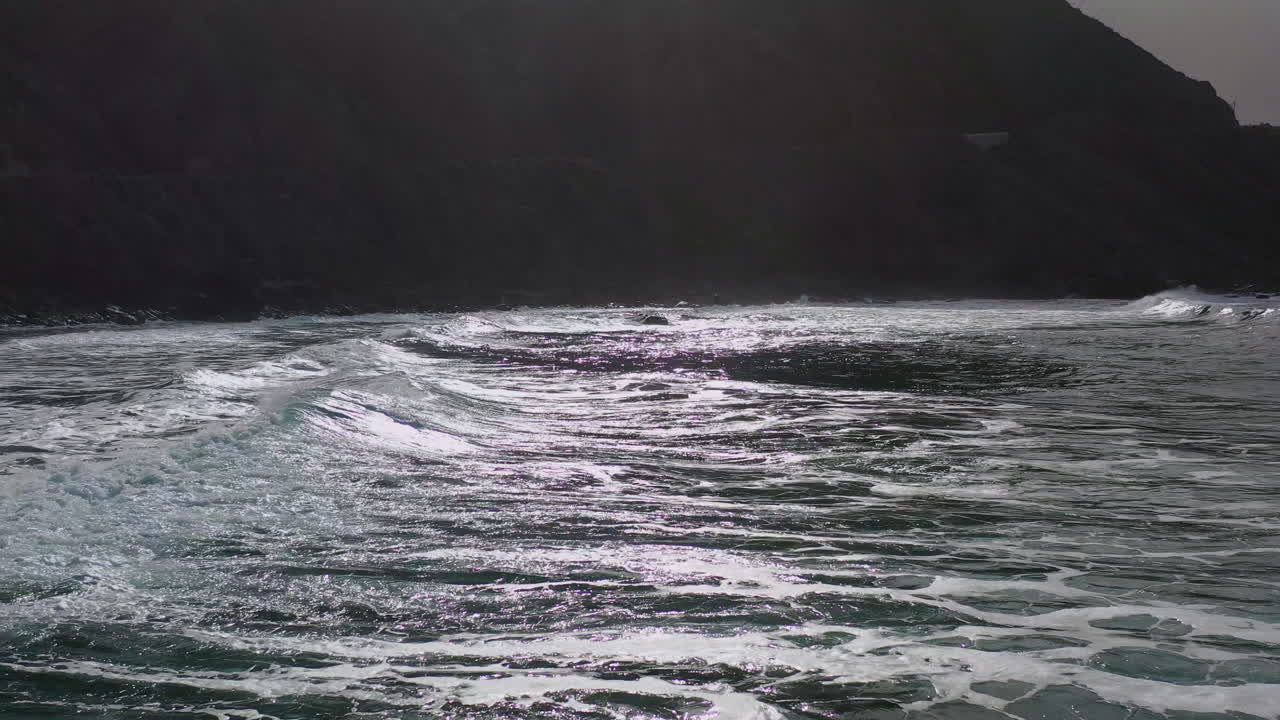 Waves washing the shallow Atlantic shoreline of the Rural de Anaga park in Tenerife, Canary Islands, Spain, with a high steep mountainous cliff in the background, forward moving 4k aerial shot.
