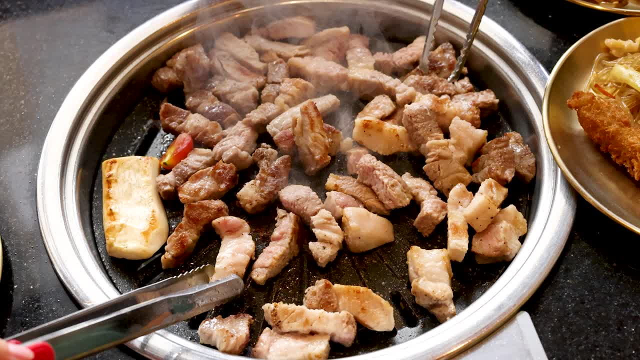 Sizzling pork belly pieces grilled on tabletop barbecue, hands using tongs, bright indoor lighting