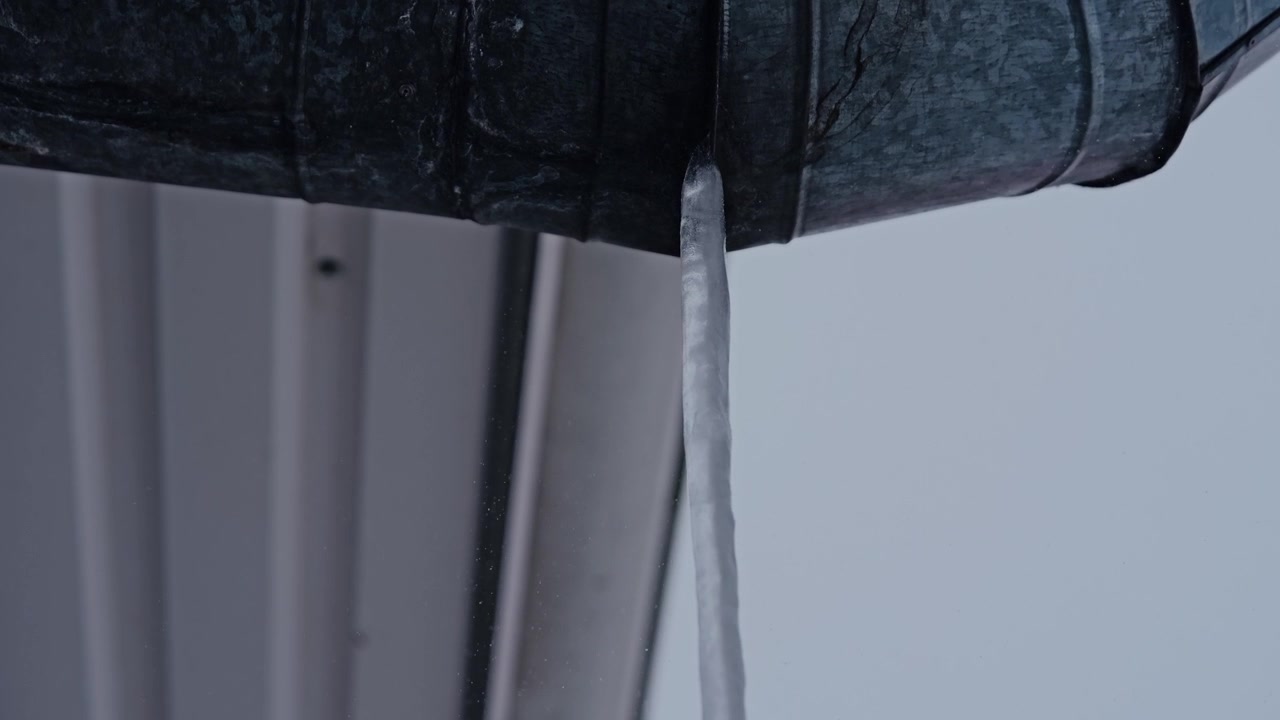 Sharp roof icicle forms in freezing weather as fine snow blows slowly past