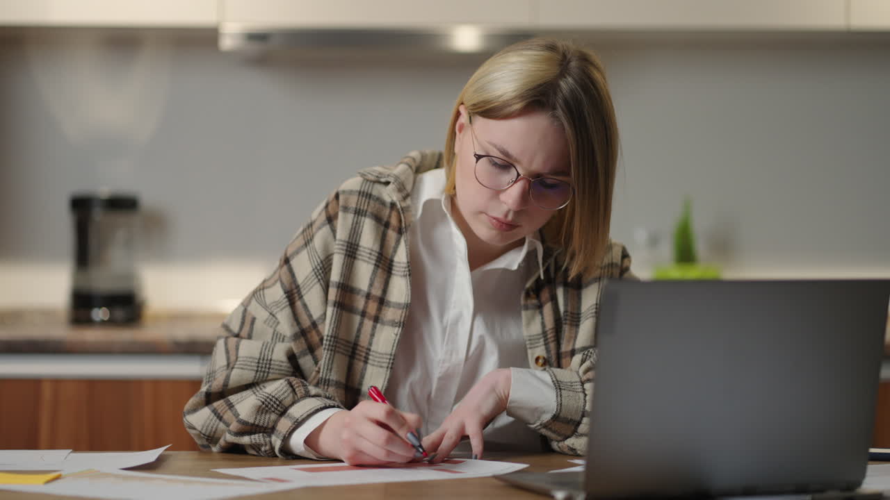 una mujer con gafas trabaja remotamente desde casa sentada en una mesa con una computadora portátil y un bolígrafo marca los datos en el gráfico.