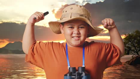 asian boy with a hat and binoculars using the magnifying glass then flexing his bicep at a lake. boy researcher examines something, travel tourism adventure, close up