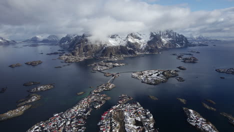 Drohnenaufnahmen-über-Henningsvær-Zeigen-Den-Atemberaubenden-Kontrast-Von-Schneebedeckten-Bergen,-Leuchtend-Blauem-Wasser-Und-Verstreuten-Inseln-Des-Lofoten-Archipels-Unter-Einem-Wolkenverhangenen-Himmel