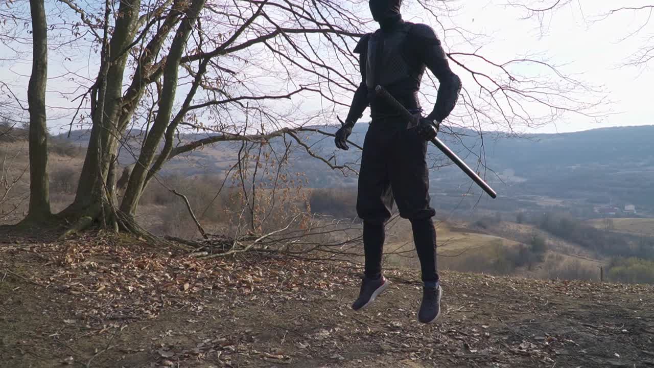 A captivating stock image featuring a stealth ninja in black, expertly unfolding and folding his katana amid a sparse forest with bare trees and rugged hills. Perfect for martial arts and action films