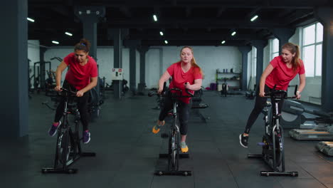 Group-of-smiling-friends-women-class-exercising,-training,-spinning-on-stationary-bike-at-modern-gym