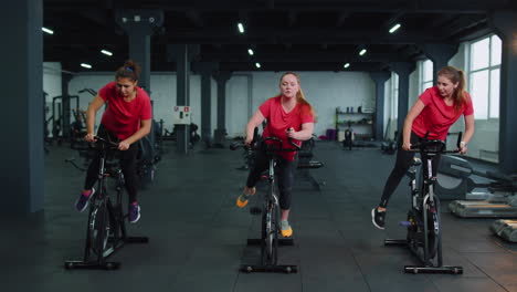 grupo de amigas sonrientes mujeres clase de ejercicio, entrenamiento, girar en bicicleta estacionaria en un gimnasio moderno