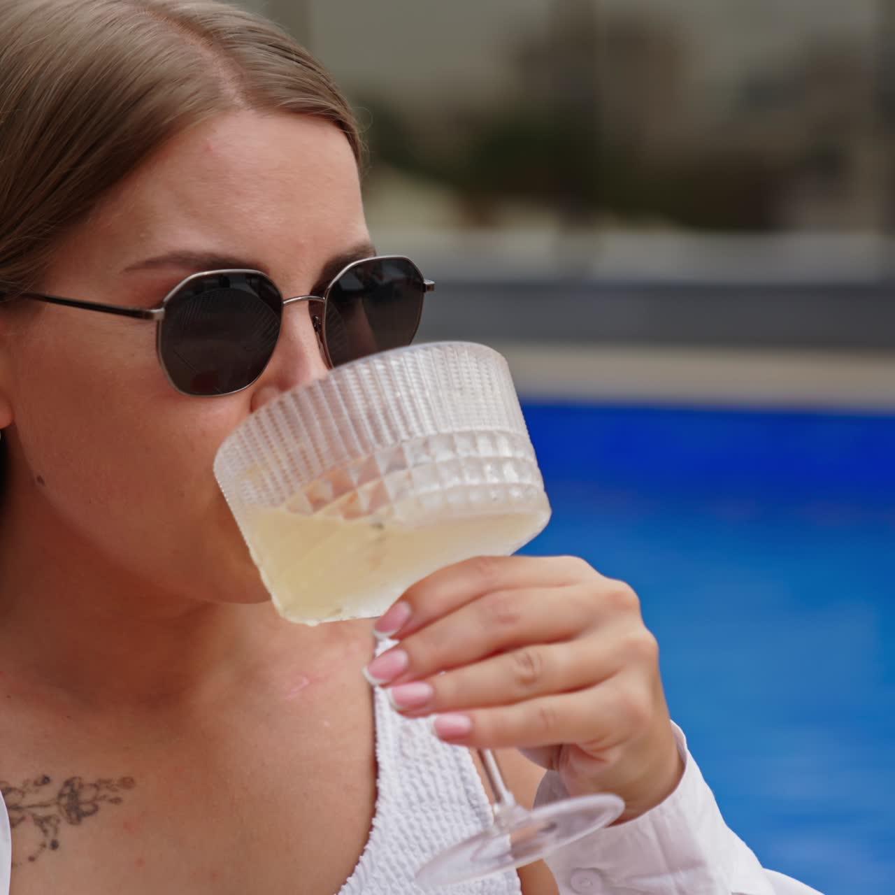 Smiling blonde girl in sunglasses and white swimsuit and shirt enjoys cocktail. Portrait of a happy lady resting at the pool