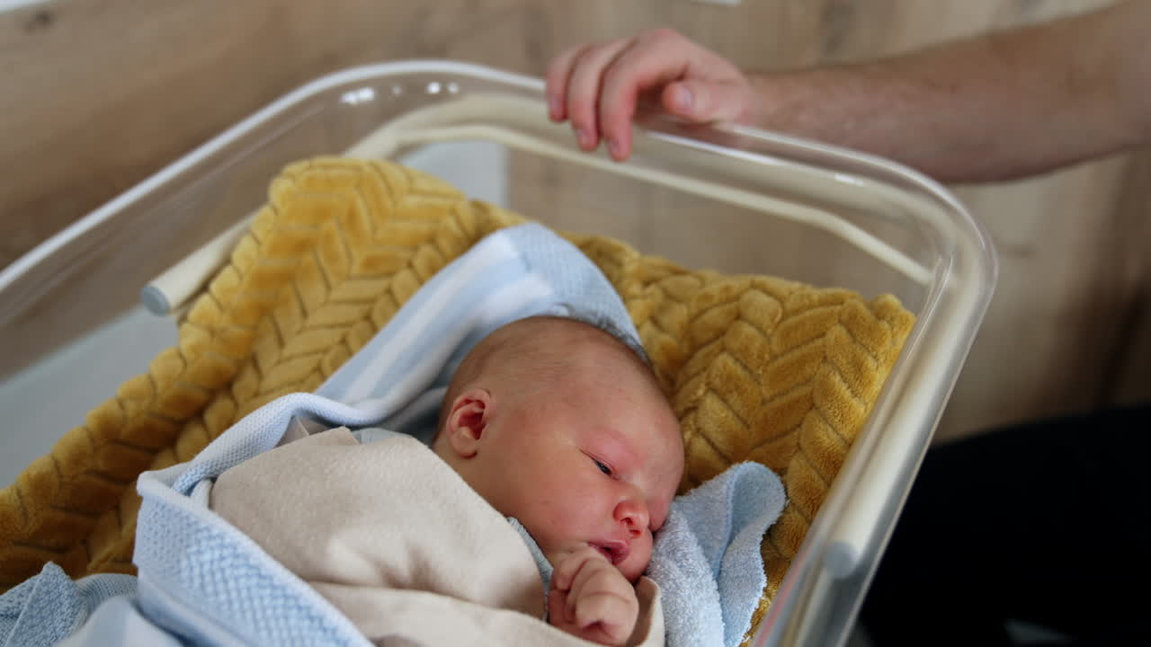 Tiny Caucasian infant in a hospital crib. Newborn baby opening his eyes slowly. Father sits beside the cradle. Top view.