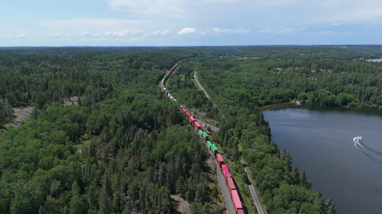 High aerial drone footage of a freight line carrying colorful shipping containers through dense forest landscape alongside a lake