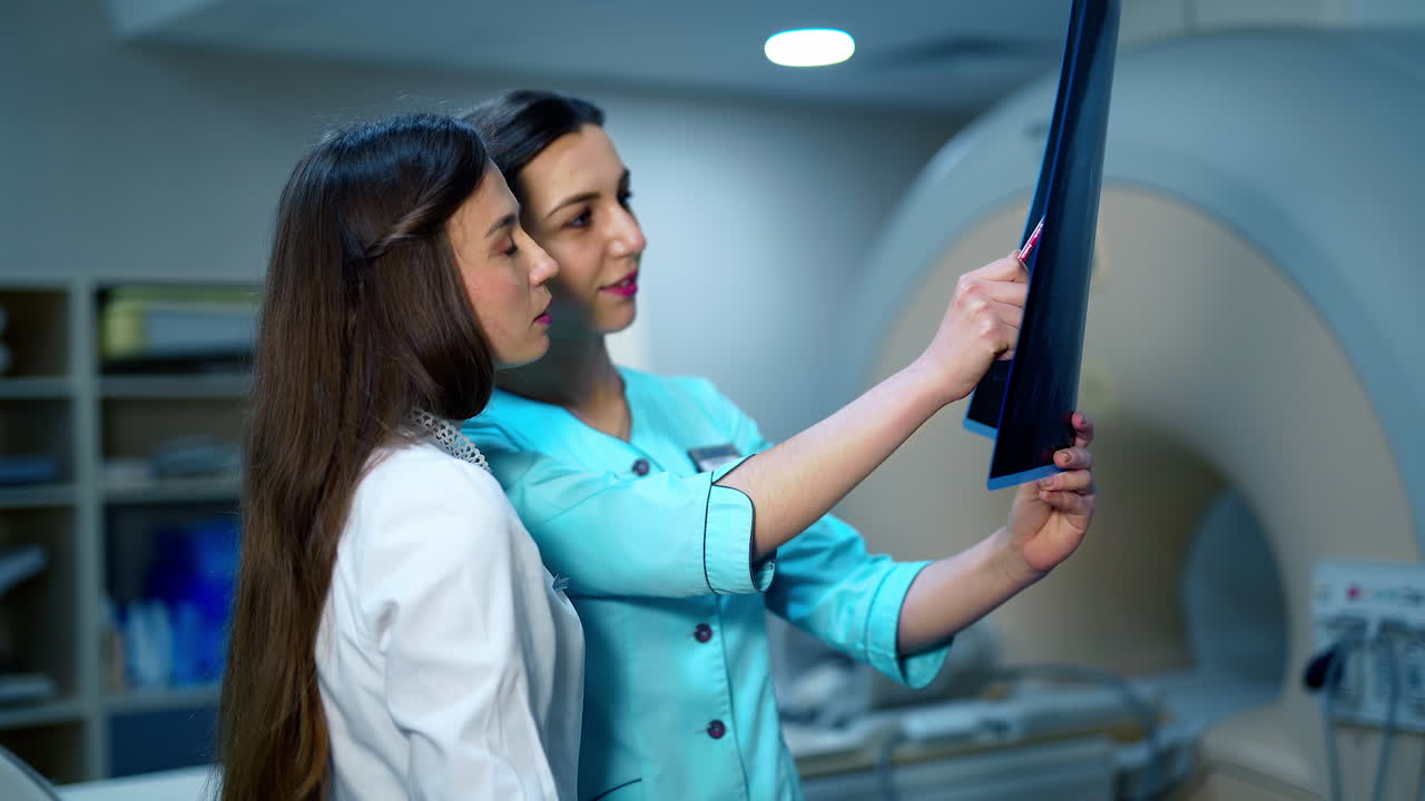 Female doctors analyzing x-ray together. Young women in medical uniform examining x-ray photo of a patient on the background of modern equipment in clinic.