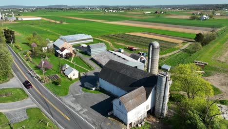 Establecimiento-De-Una-Toma-De-Drones-De-Un-Automóvil-Conduciendo-Por-Un-Camino-Rural-Al-Lado-De-Una-Granja-Con-Establo,-Almacenamiento-De-Silos-Y-Campos-Coloridos-En-Primavera