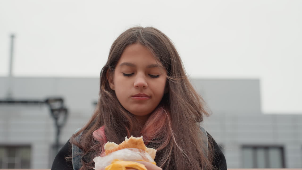teen student on bench eating sandwich wrapped in yellow paper wearing scarf and coat gazing thoughtful against modern campus architecture overcast sky candid portrait capturing quiet hunger