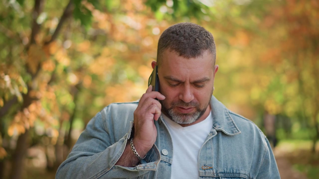 Nærbilde av mann som snakker i telefonen i en park med høstløvverk i bakgrunnen. Mannen har på seg dongerijakke og hvit skjorte, holder en svart smarttelefon til øret. Mykt naturlig lys lyser opp scenen.