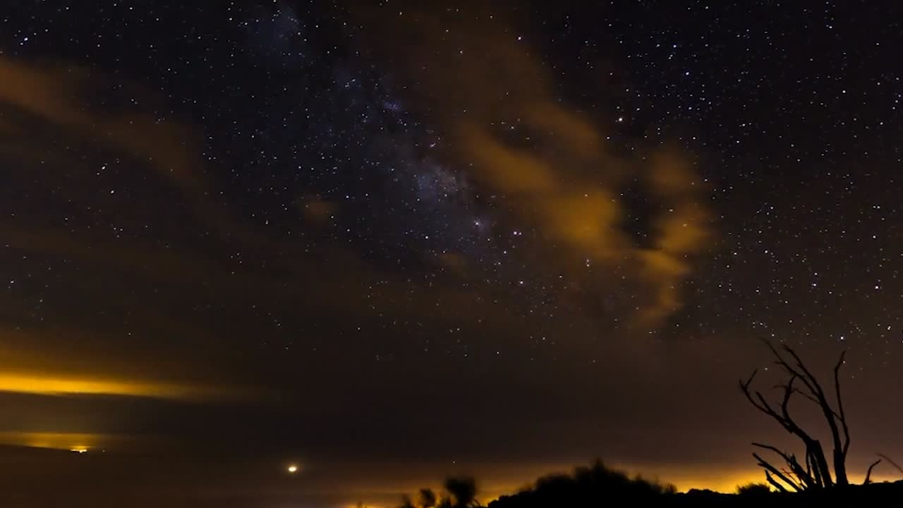 Time-lapse-opptak av en stjerneklar nattehimmel som viser det svake, skyete båndet av Melkeveigalaksen som strekker seg over horisonten. Tynne skyer gløder med varmt oransje lys fra en fjern by.