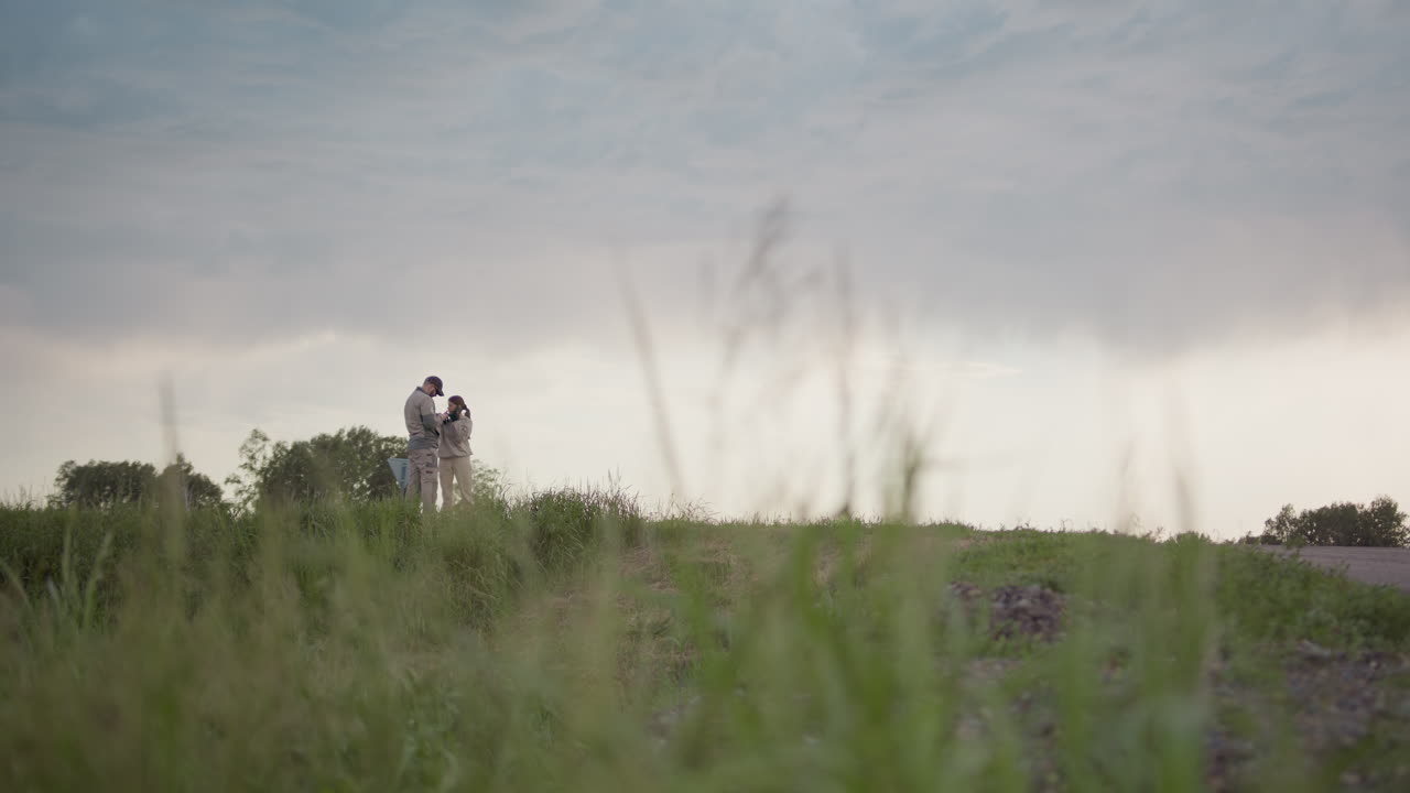 Two people stand on grassy roadside under cloudy sky, woman talks on phone while man checks handheld device, black car drives by behind them
