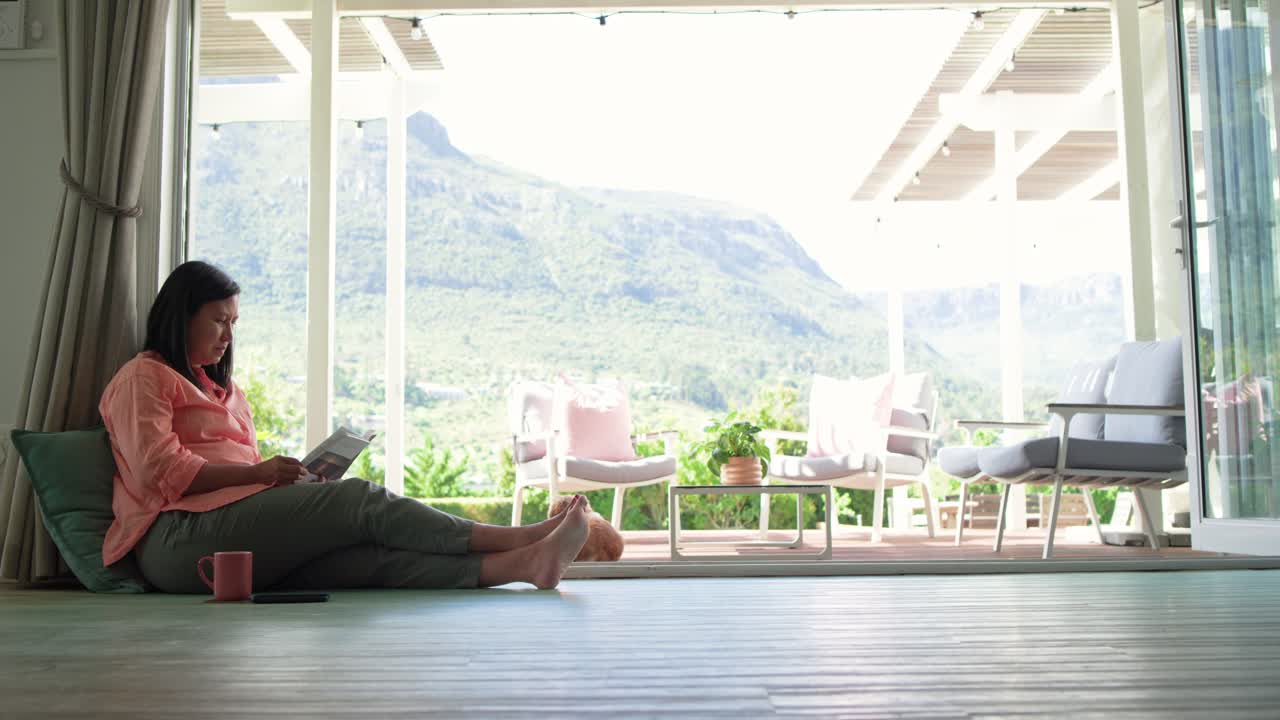 Femme asiatique dessinée par le soleil, assise par terre près d’une vitre, lisant un livre, tendant la main vers une tablette, tasse, espace réservé