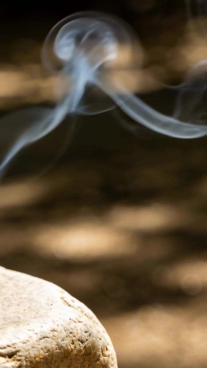 Vertical video: Rising thin blue-gray smoke curling and drifting over soil near rock from ember