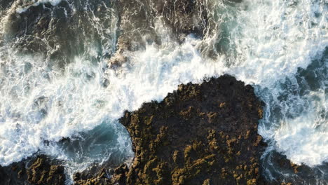 Vista-De-Arriba-Hacia-Abajo-De-Las-Olas-Rompiendo-En-Las-Rocas-Volcánicas-De-La-Playa-De-Arena-En-Oahu,-Hawaii