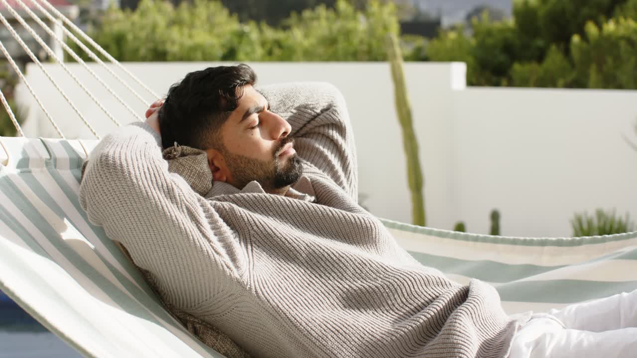 Uomo indiano che inspira, sdraiato e che respira su un'amaca a righe su una terrazza con cuscino per rilassarsi