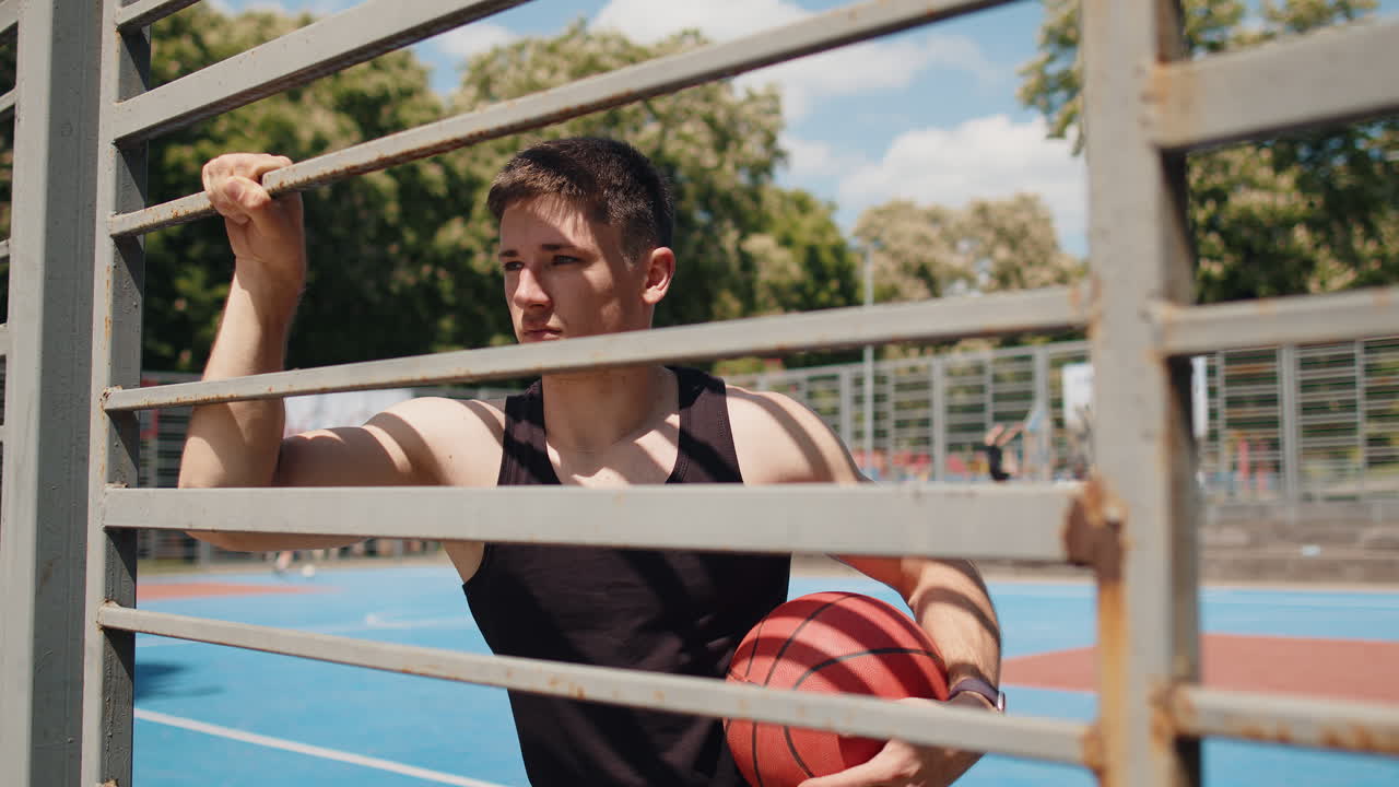 Portrait of athletic serious man playing basketball posing alone stands behind grid on urban court