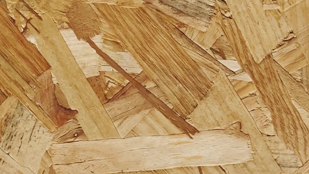 Detailní záběr na OSB desku s vrstvenými dřevěnými vlákny, hrubou texturou lisovaných třísek, teplými béžovými tóny a detaily povrchu stavebního materiálu.