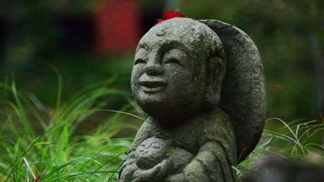 a small jizo staute with a red leaf on its head, staying in lush green grass