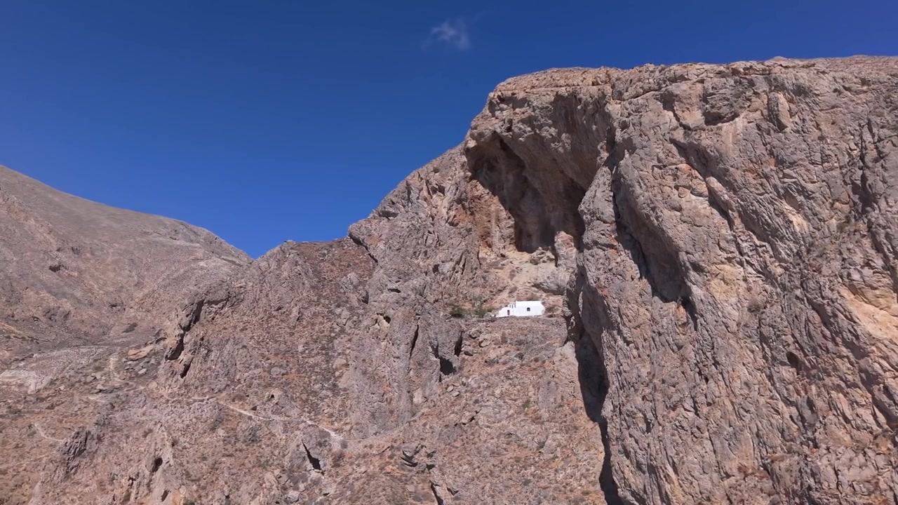 Ripresa aerea che rivela una cappella bianca isolata arroccata sul fianco roccioso della montagna sopra Perissa. Il filmato cattura il contrasto tra il terreno roccioso e il profondo Mar Egeo blu sottostante